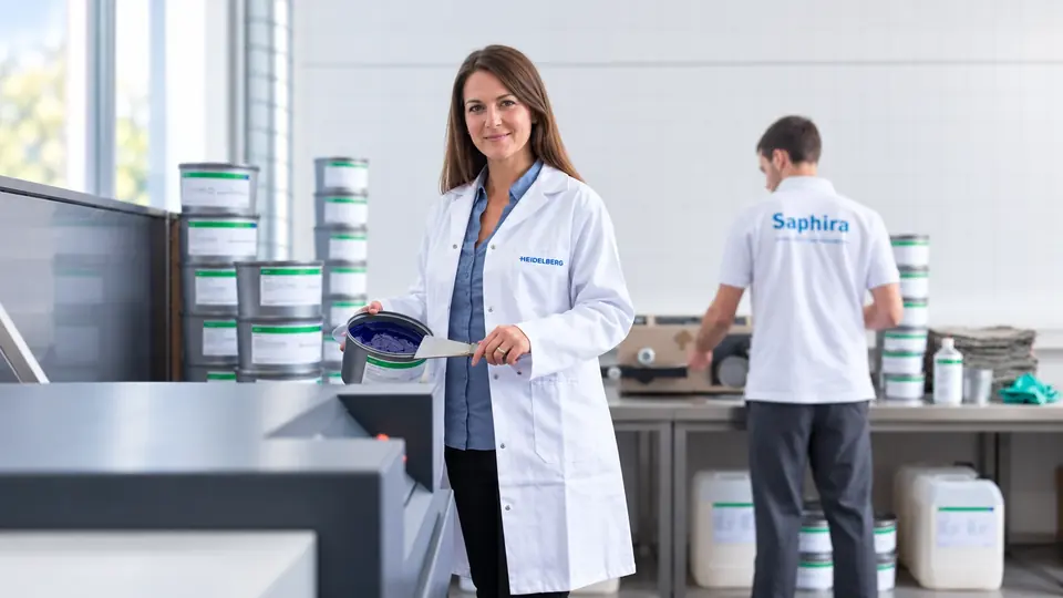 Technicienne en blouse Heidelberg tenant un pot d’encre offset, devant un stock d’encres et de produits Saphira